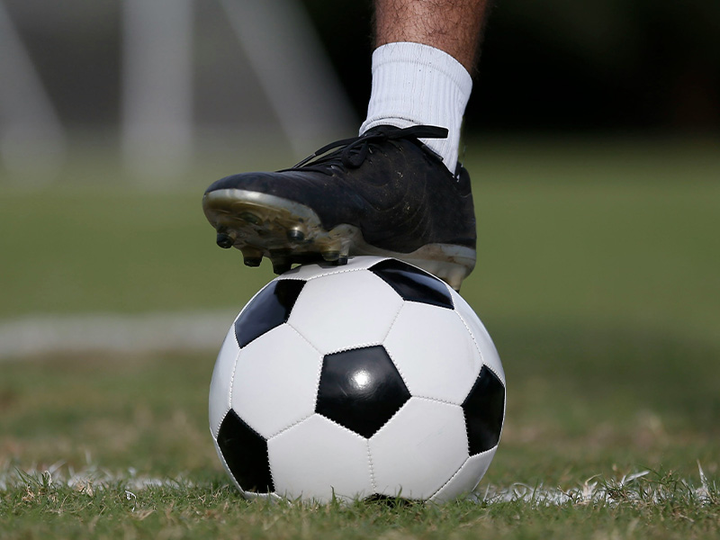 A soccer player’s cleat rests on top of a black-and-white soccer ball on a grassy field.