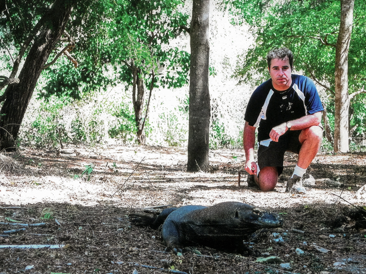 Peter Alexander kneels on a wooded forest floor, observing a Komodo dragon resting on the ground nearby.