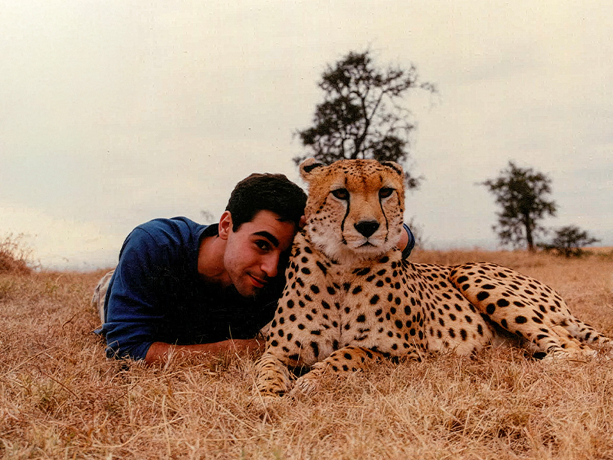Peter Alexander lies in dry grass beside a resting cheetah, his head close to the animal’s shoulder.